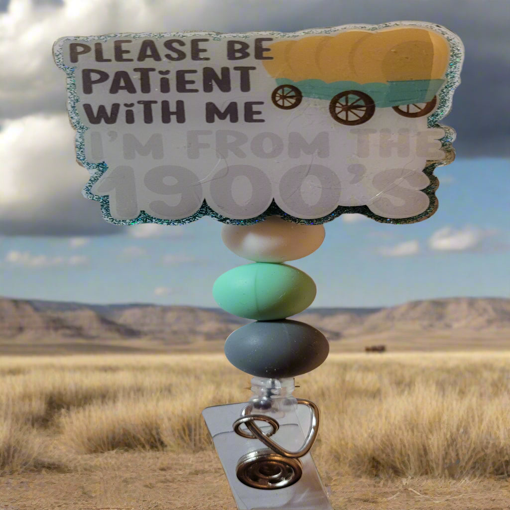 Get ready to travel back in time with this playful Badge Reel inspired by the 1900's! The front features a covered wagon, a nod to the Oregon Trail, and the catchy phrase "Please Be Patient With Me I'm From The 1900's". The back is a fun blue green glitter and is completed with 3 matching silicone beads.