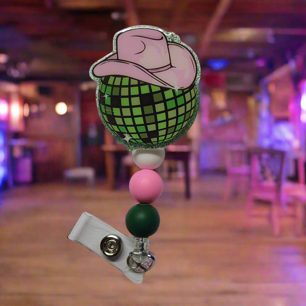 Every cowgirl needs a hat. This acrylic badge reel showcases a classic cowgirl hat in the color pink atop a green disco ball all ready for a night of dancing at the local honkey tonk. A green glitter back and 3 silicone beads finish the look.