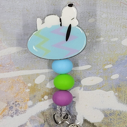 Everyone's Favorite Beagle has replaced is dog house with an Easter Egg as he takes his daily nap. Laying on a light blue egg with pink and green zig zag stripes on a clear glitter background and adorned with three perfectly matching silicone beads, adding a charming touch to this delightful accessory. A must-have for all animal enthusiasts!