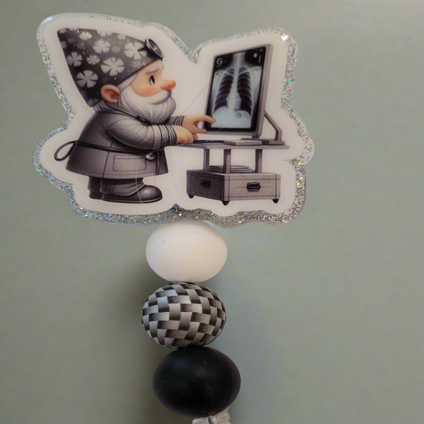 Introducing the new Gnome X-Ray Tech Badge Reel, part of our latest collection of Medical Badge Reels available now. Featuring a cute Gnome examining an X-Ray dressed in shades of gray and a silver glitter base adorned with silver black & white 3d visual bead and s more color coordinating beads, this Badge Reel is sure to catch the eye.