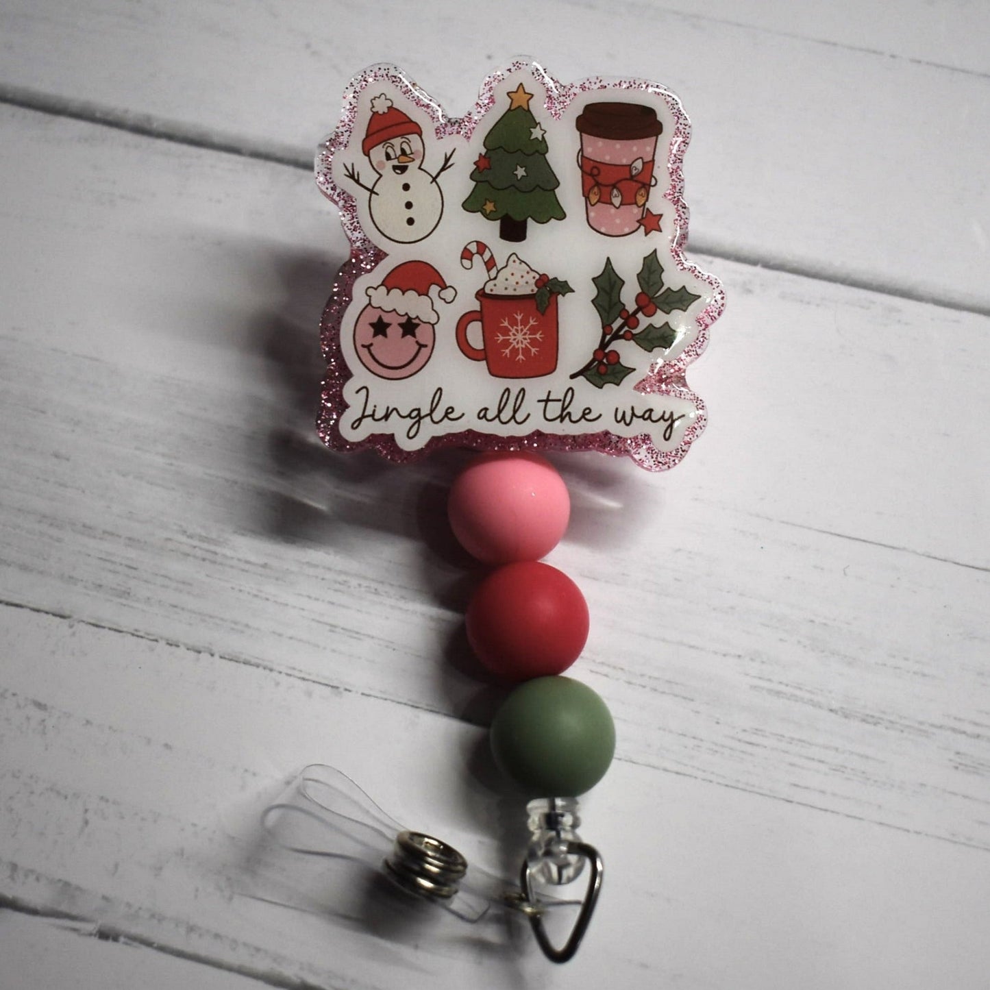 Featuring a pink glitter background atop a white base and six images of classic Christmas motifs, this Badge Reel Jingle All The Way is further adorned with green, red, and pink accent beads, creating a unified look.