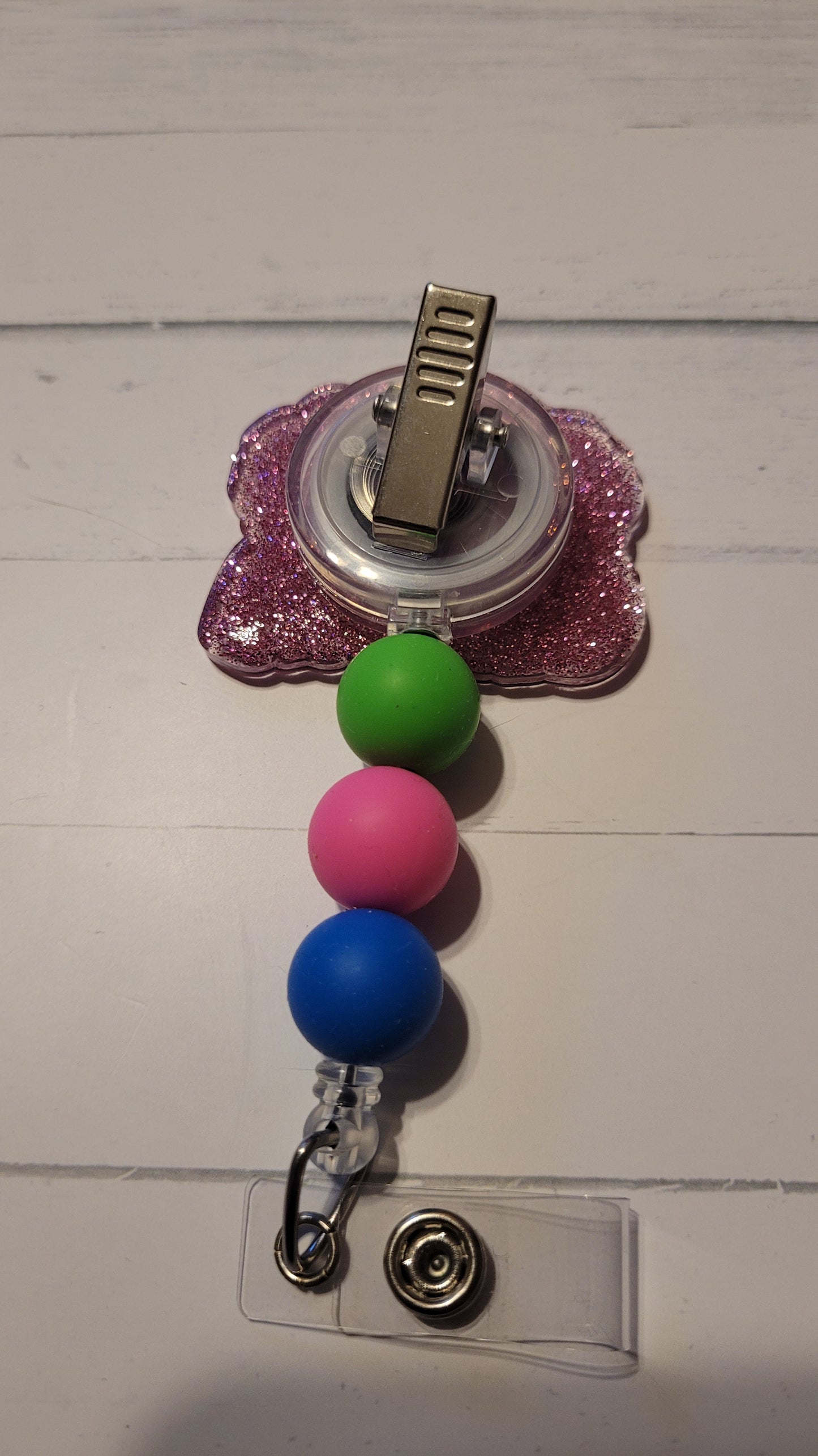 The Badge Reel Ohana showcases our happy little alien indulging in his preferred beverages on a palm tree covered beach. The elegant design is enhanced by a sparkling purplish glitter base and three color-coordinated beads.
