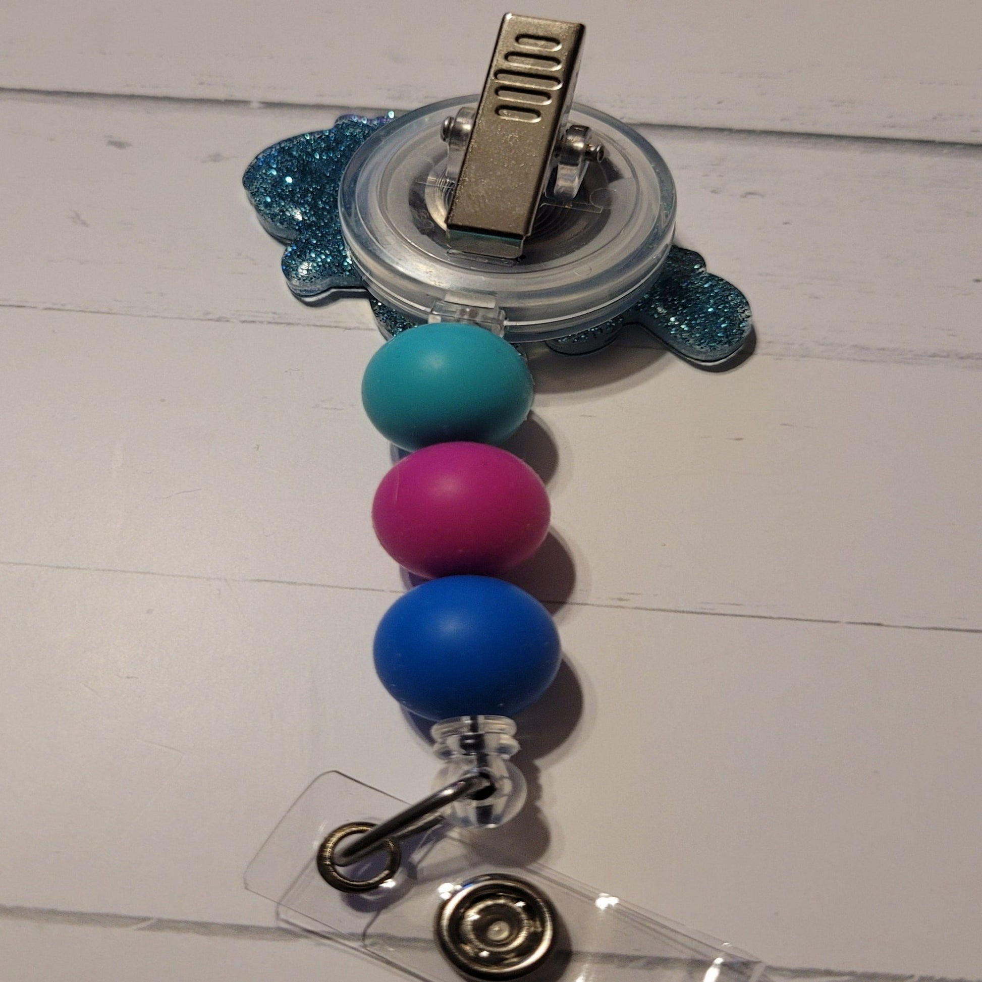 This delightful badge reel features a cheerful blue turtle with a bright pink rose atop its head. It also boasts a sparkly blue background and three cute matching silicone beads for added charm. An absolute must-have for all animal lovers!