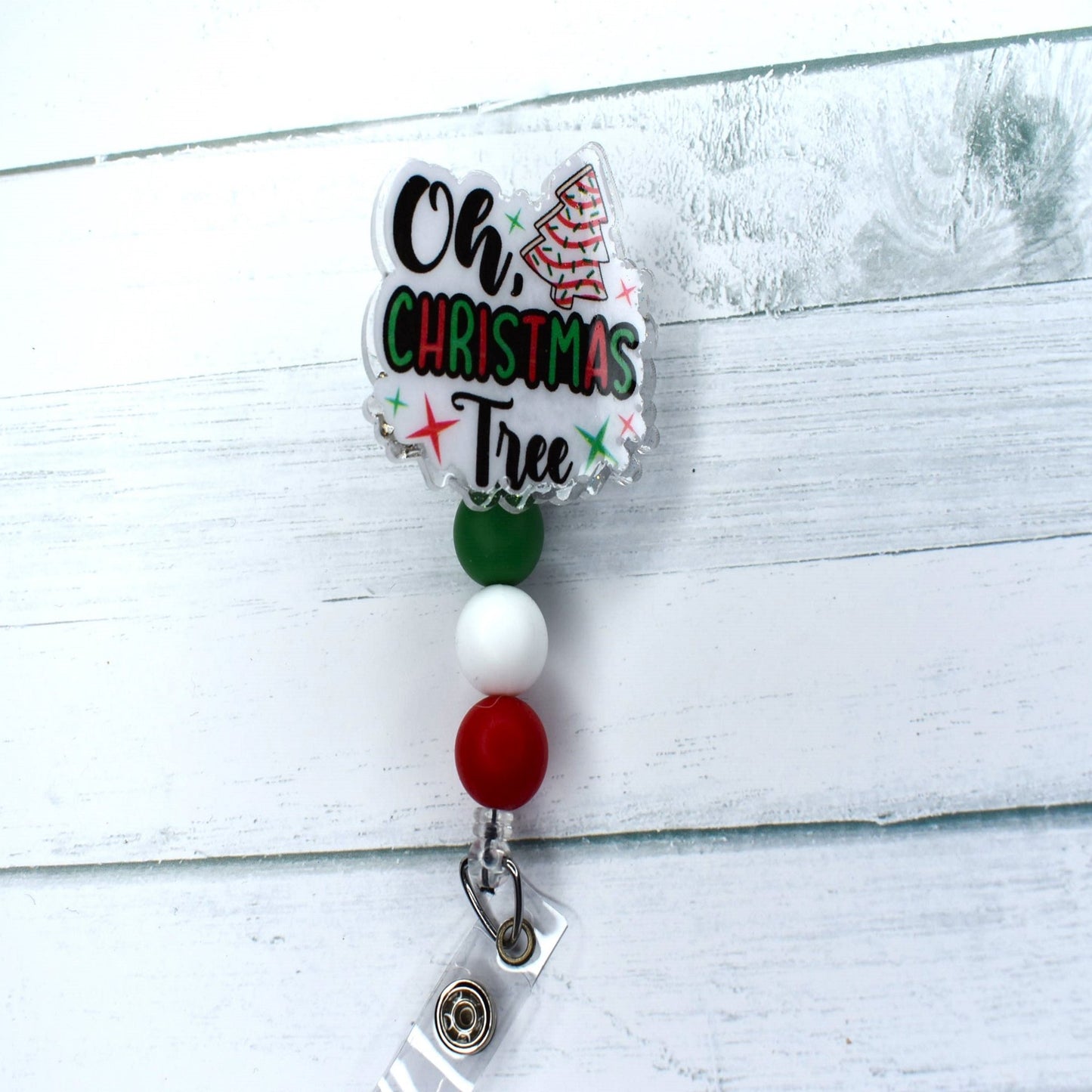 This acrylic badge reel proudly displays a classic holiday treat - the Christmas Tree Snack cake. A white background and festive-hued silicone beads complete this seasonal delight, which is tastefully adorned with the words "Oh Christmas Tree".