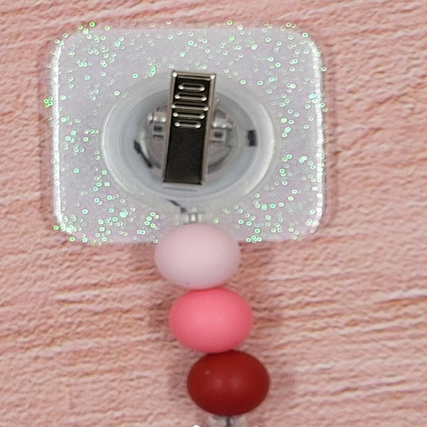 This 2" x 2" Badge Reel features a Valentines themed Alphabet Chart in a combination of pink and red hues. Adding to its festive appeal is an iridescent glitter base, accompanied by three silicone beads in coordinated colors. Elevating your style for the holiday season, this is a must-have accessory