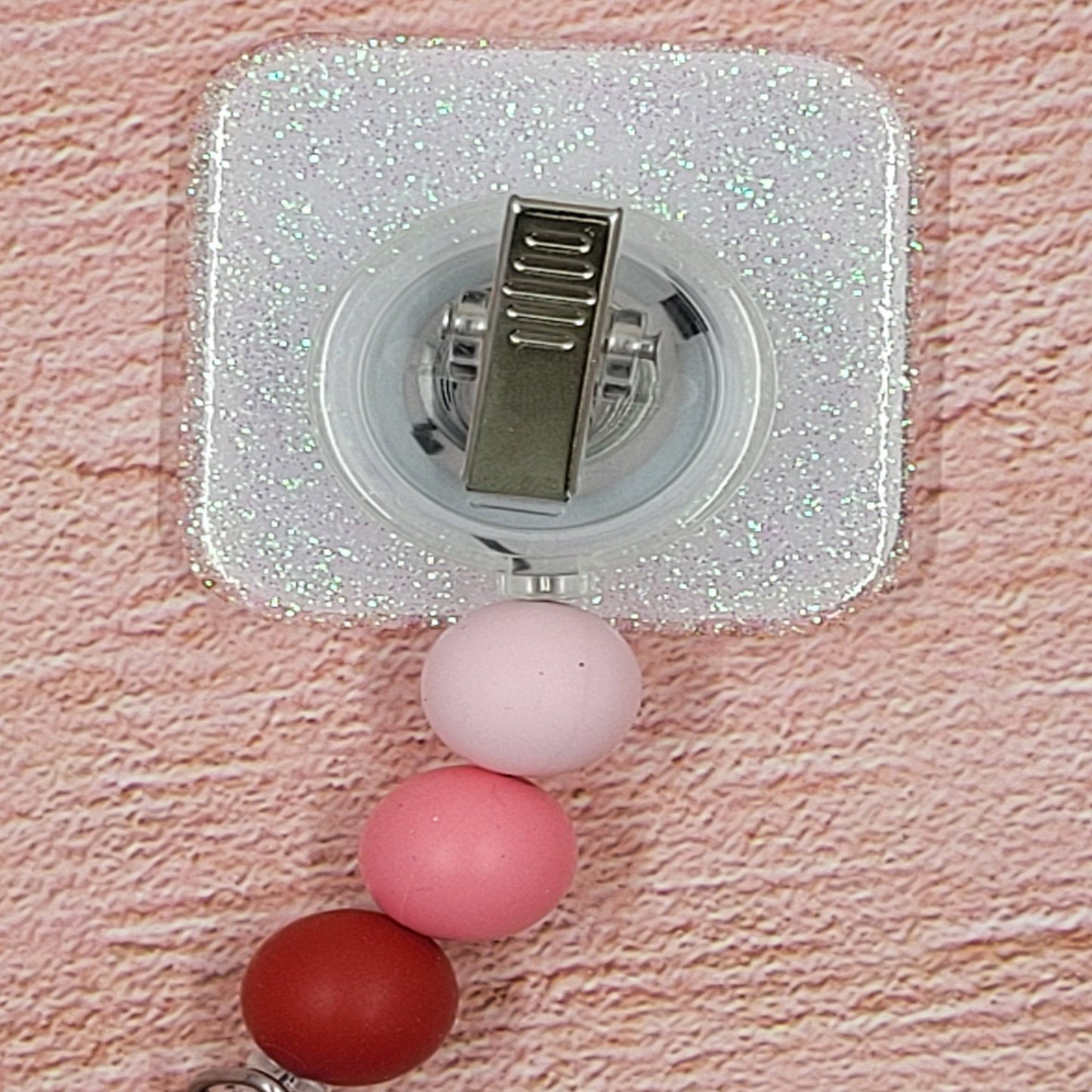 This 2" x 2" Badge Reel features a delightful combination of pink and red hues, adorned with the charming phrase "Hugs Kisses & Valentine Wishes". Adding to its festive appeal is an iridescent glitter base, accompanied by three silicone beads in coordinated colors. Elevating your style for the holiday season, this is a must-have accessory.