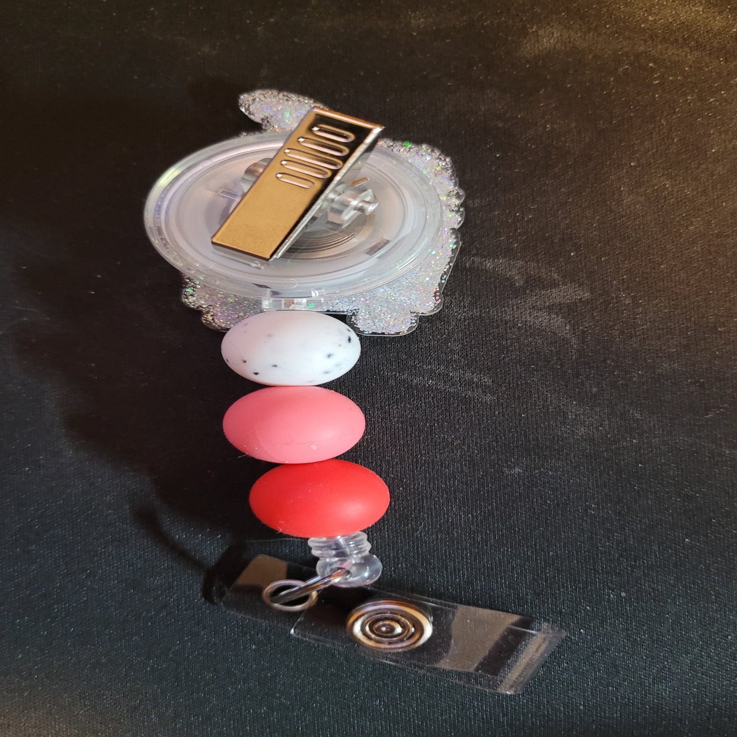 The Badge Reel Valentine Love Gnome Wagon features a cute gnome in a pink knit hat surrounded by flowers as he rides in a wagon on a mission to spread love. With a clear glitter base and 3 color matching silicone accent beads, this badge reel is sure to please.