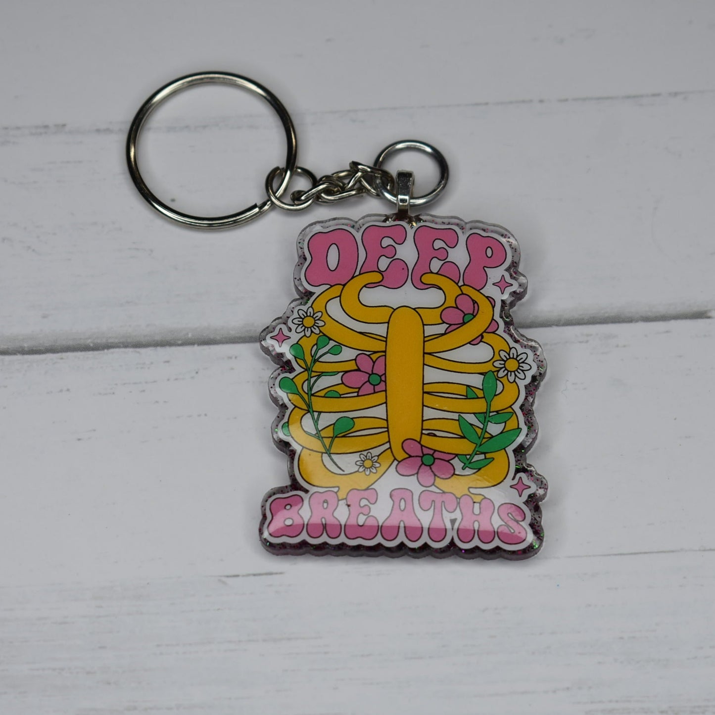 Feeling overwhelmed? Take a moment to relax and just breathe. The Keychain Deep Breaths serves as a helpful reminder to pause, take a deep breath, and refocus. With its sturdy acrylic resin material, it boasts a stylish purple glitter backing and a motivational Deep Breaths design on the front.