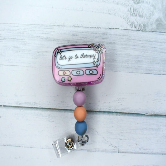 This acrylic badge reel serves as a reminder that mental wellness is important. Let's Go To Therapy is the message here, available in either pink or purple with coordinating silicone beadwork.