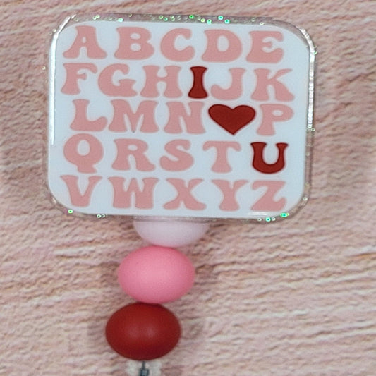 This 2" x 2" Badge Reel features a Valentines themed Alphabet Chart in a combination of pink and red hues. Adding to its festive appeal is an iridescent glitter base, accompanied by three silicone beads in coordinated colors. Elevating your style for the holiday season, this is a must-have accessory