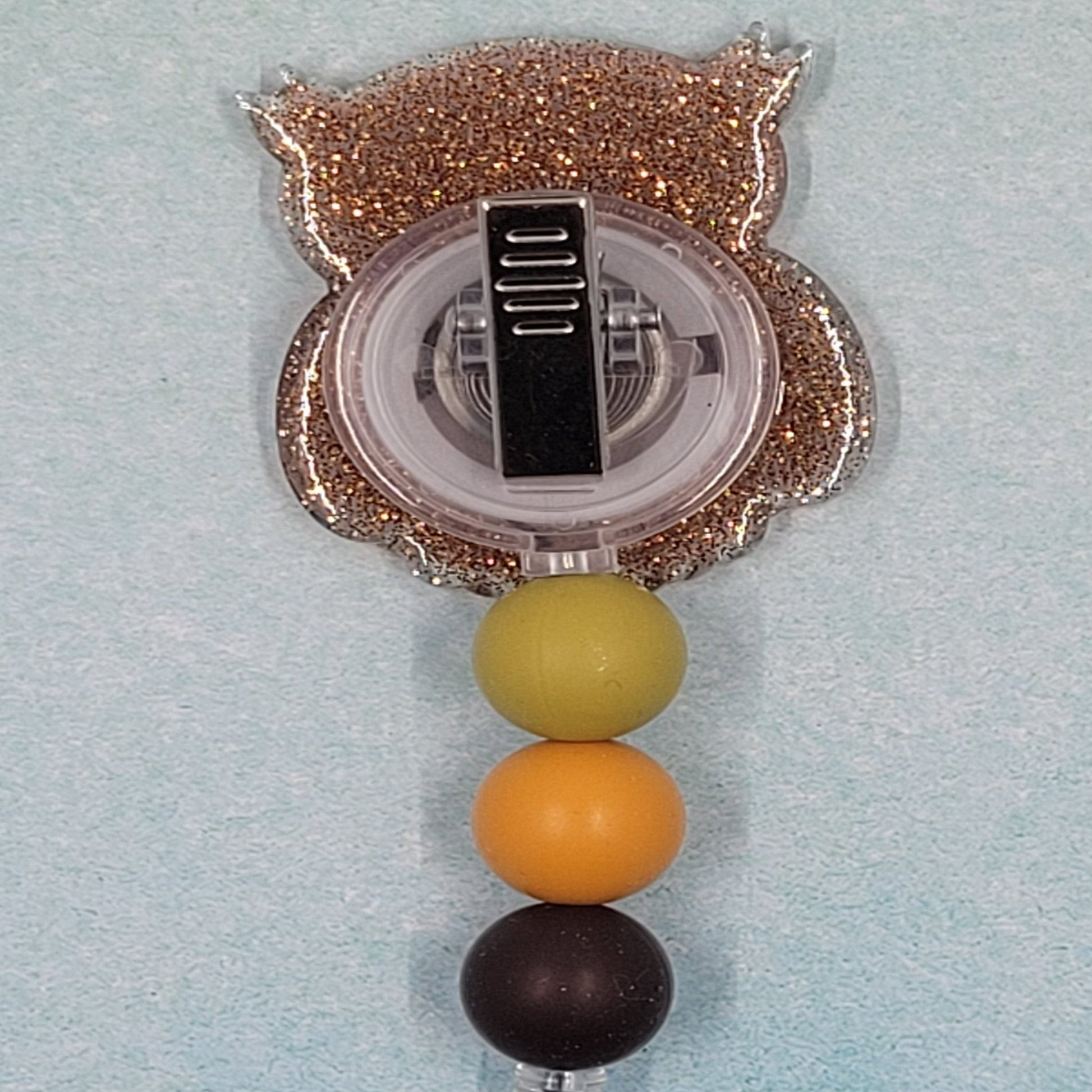 This Badge Reel features a Wise Owl design paired with a shiny brown glitter background. The adorable owl is complemented by three color-coordinated silicone beads for an extra touch of charm. Sure to be a hit with any animal lover.