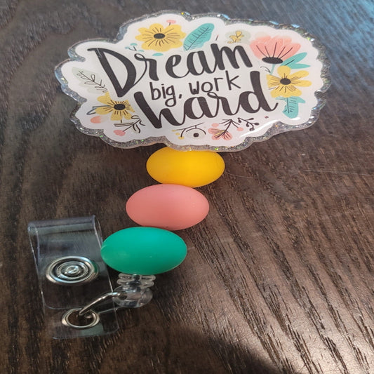 Dream Big Work Hard is our newest inspirational badge reel. Those words are centered on the front and surrounded by various flowers. Decked With a sparkling clear glitter back and embellished with 3 silicone beads on the cord